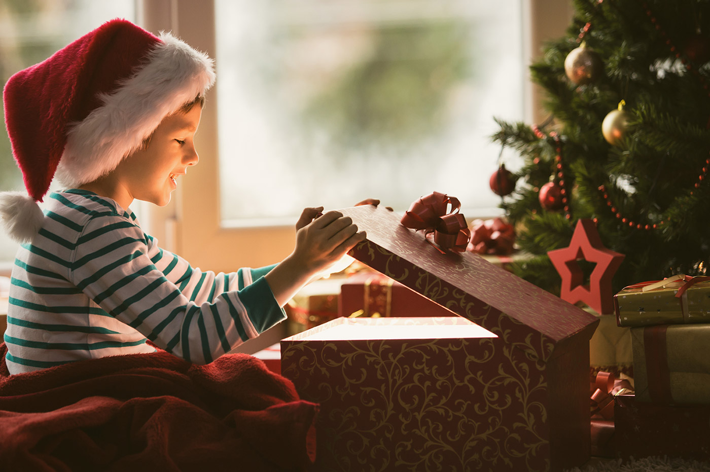 A little boy in pajamas and a Santa hat, opening up a Christmas present by the tree.