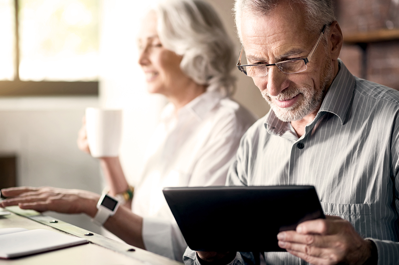 An older woman drinking a cup of coffee and an older man looking at a tablet. 